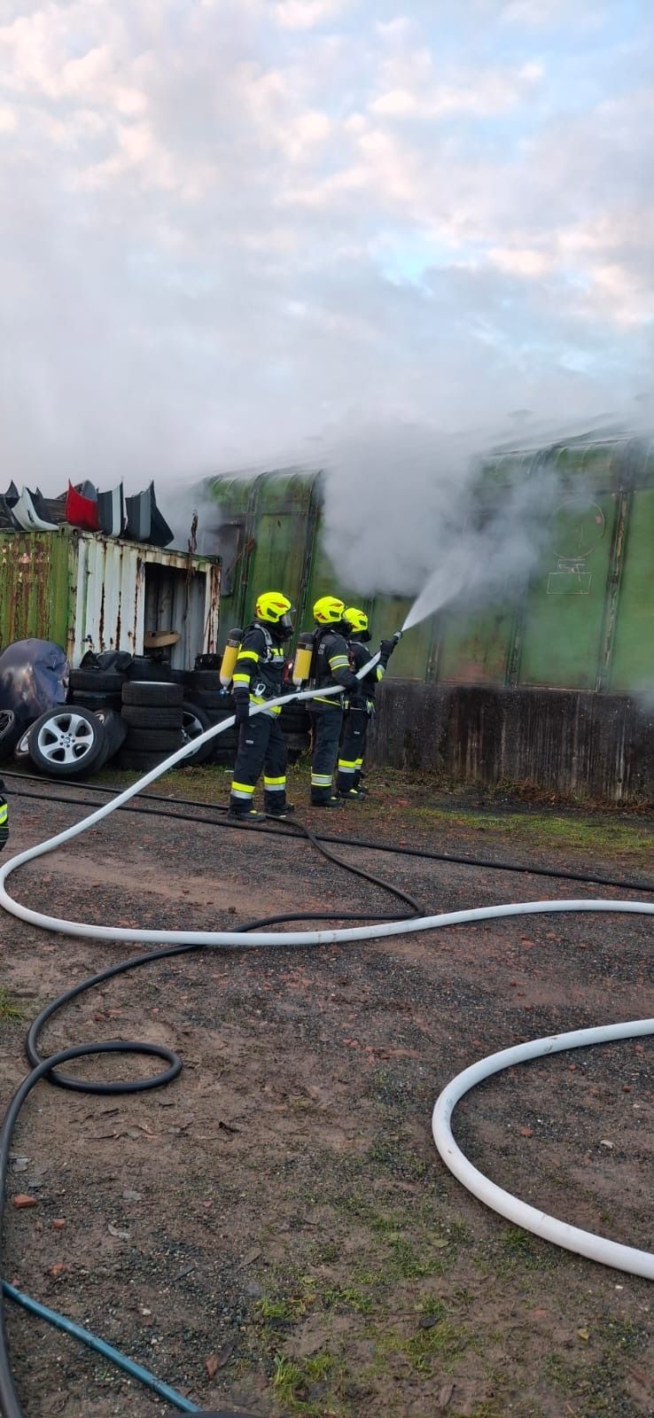 Drei Feuerwehrleute sprühen Wasser aus einem Schlauch, um ein Feuer in einem Waggon zu löschen. In der Nähe brennt ein Reifenstapel.