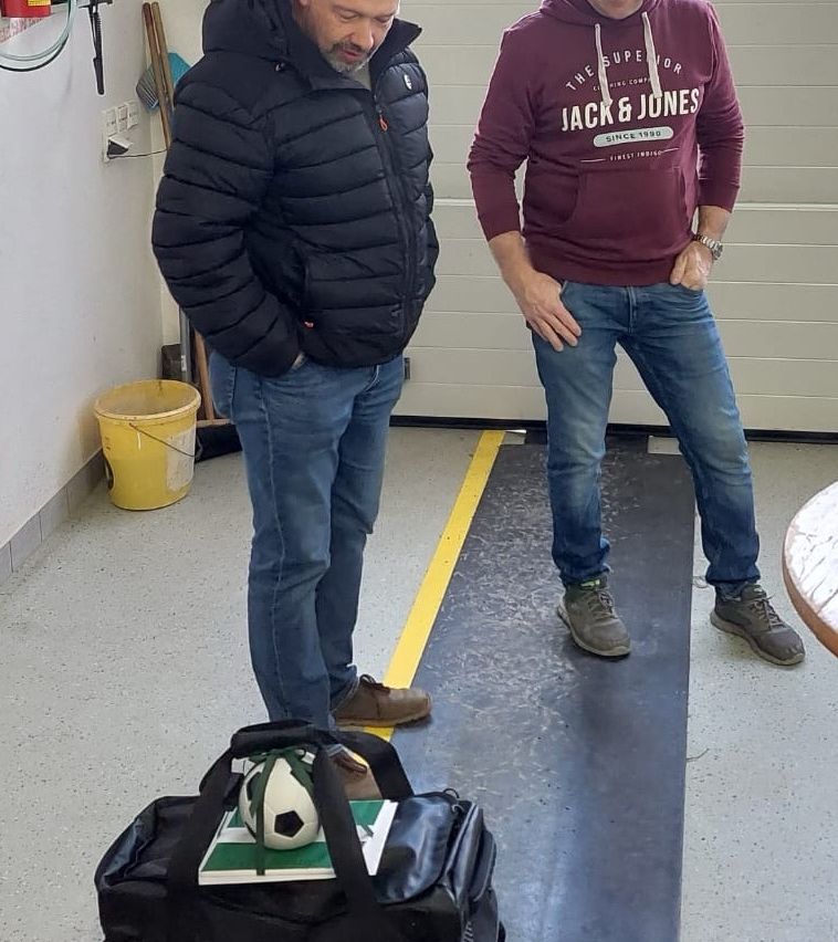 Zwei Männer stehen in einem Raum mit einem Fußball auf einer Tasche, einer trägt eine schwarze Jacke, der andere ein weinrotes Sweatshirt. Ein gelber Eimer und ein Tisch sind in der Nähe.