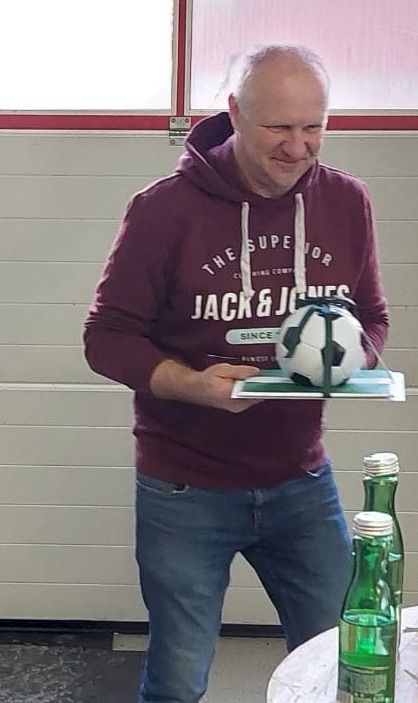 Ein Mann in einer weinroten Kapuzenjacke steht vor einem Tisch mit zwei grünen Flaschen und hält lächelnd einen Fußball in der Hand.