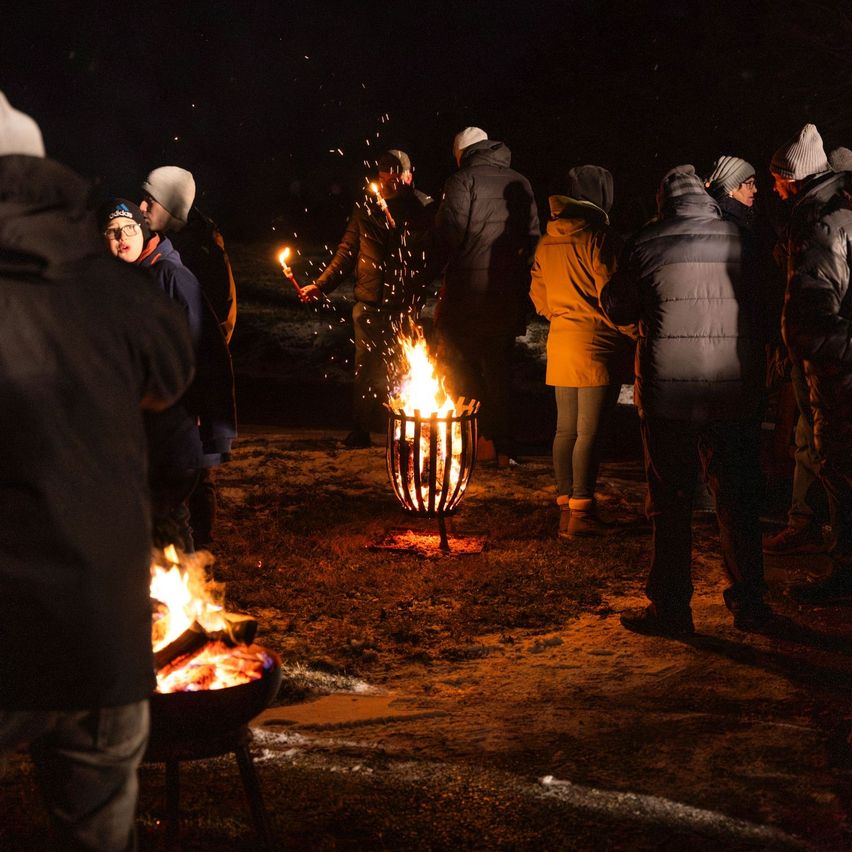 Eine Gruppe von Menschen versammelt sich bei Nacht um ein Lagerfeuer, einige halten brennende Fackeln. Sie sind warm gekleidet, tragen Winterkleidung und Mützen.
