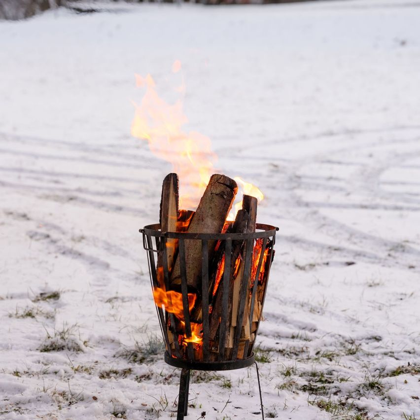 Ein metallner Feuerkorb mit brennendem Holz steht auf einem verschneiten Feld mit Reifenspuren.