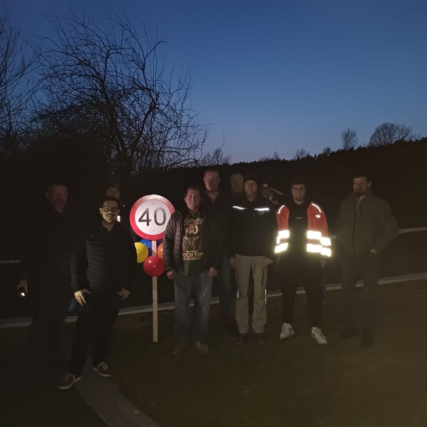 Eine Gruppe von Männern steht bei einem 40 km/h Geschwindigkeitslimit-Schild und posiert für ein Foto bei Nacht. Sie tragen Jacken und einige haben reflektierende Westen.