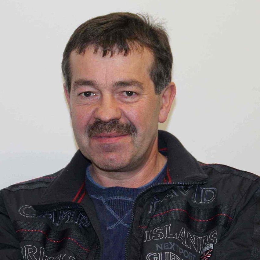 Ein Mann mit einem Schnurrbart, der eine schwarze Jacke mit weißen und roten Nähten trägt, er schaut in die Kamera und lächelt.