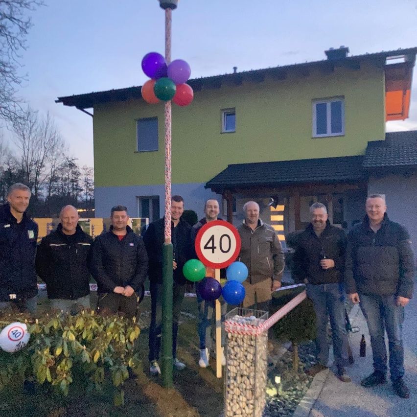 Eine Gruppe von Männern steht vor einem Haus mit einem 40er-Schild, umgeben von Luftballons und einem Korb mit Steinen.