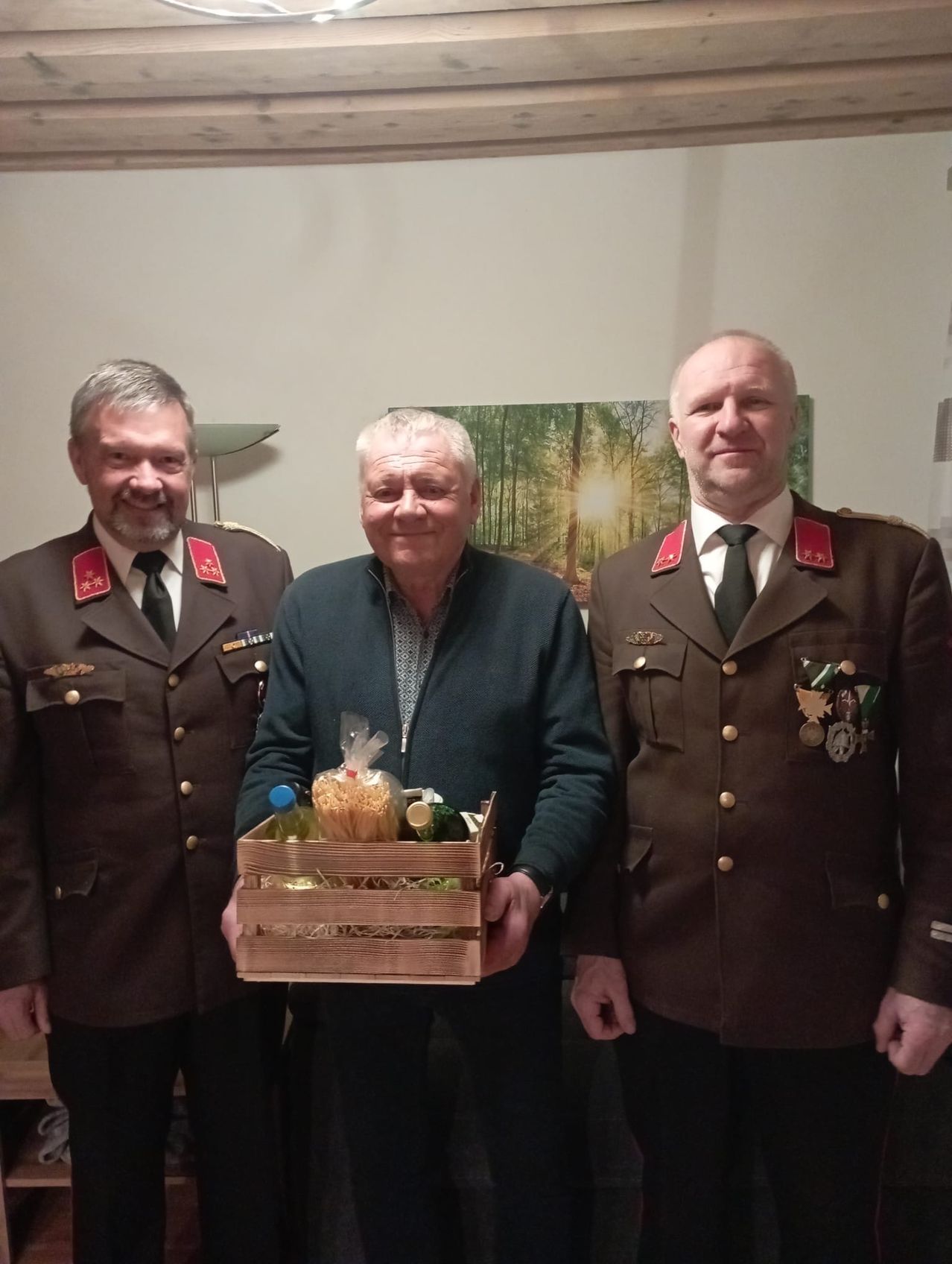 Drei Männer in Militäruniformen stehen eng beieinander und lächeln. Der Mann in der Mitte hält eine Holzkiste mit Lebensmitteln. Hinter ihnen ist eine weiße Wand mit einem Gemälde.