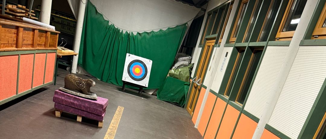 Eine Inneneinrichtung mit braunem Boden, orangefarbener Wand und Holzbalken. Ein schwarzes Objekt mit der Nummer 3 steht auf dem Boden. Eine Holzstruktur mit Ziel befindet sich links.