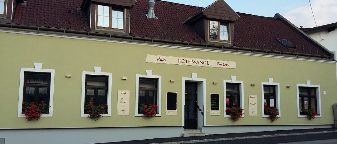 Ein gelbes Gebäude mit braunem Dach, weißer Einfassung und roten Blumen in Kisten. Es trägt den Namen Rothwangl Bäckerei über dem Eingang.