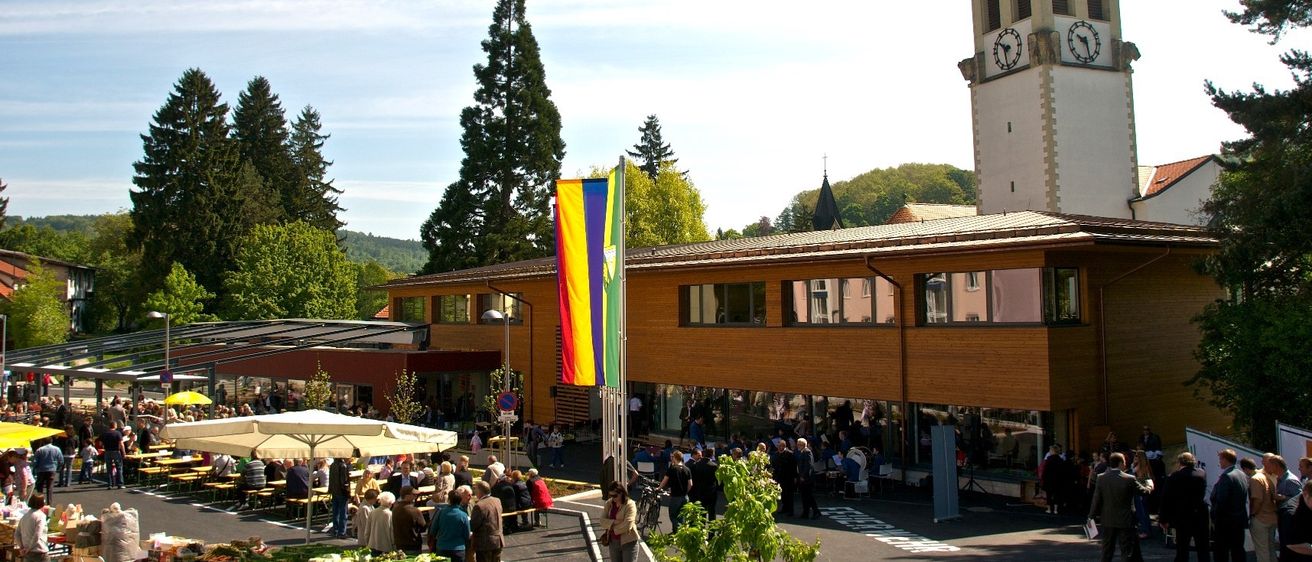 Eine Marktszene mit vielen Menschen, einem Holzgebäude, einem Glockenturm und einer Regenbogenflagge. Menschen sitzen unter Sonnenschirmen und an Tischen, während andere im Vordergrund gehen.