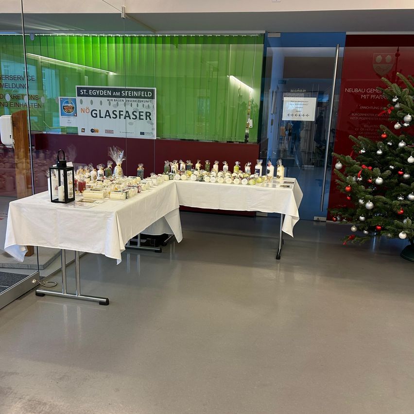 Two tables with white tablecloths hold numerous wrapped items. A Christmas tree stands beside the tables. A banner reads 'St. Eygyden am Steinfeld' and 'No Glasfaser'. A glass door shows a reflection of a person.