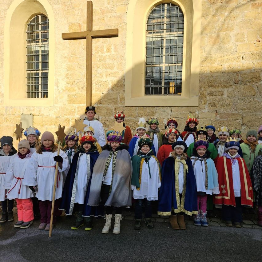 Eine Gruppe von Kindern in verschiedenen Kostümen, einige mit einem Stab, stehen vor einem Kirchengebäude mit einem Kreuz an der Wand.