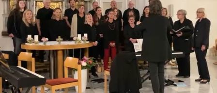 Ein Chor in Schwarz singt in einer Kirche, geleitet von einem Dirigenten, mit einem Tisch und Blumen im Hintergrund.