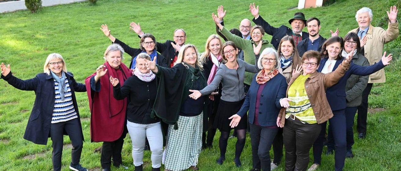 Eine Gruppe von Erwachsenen, darunter Männer und Frauen, stehen auf einer Wiese und lächeln und winken mit ihren Händen, wahrscheinlich für ein Foto.