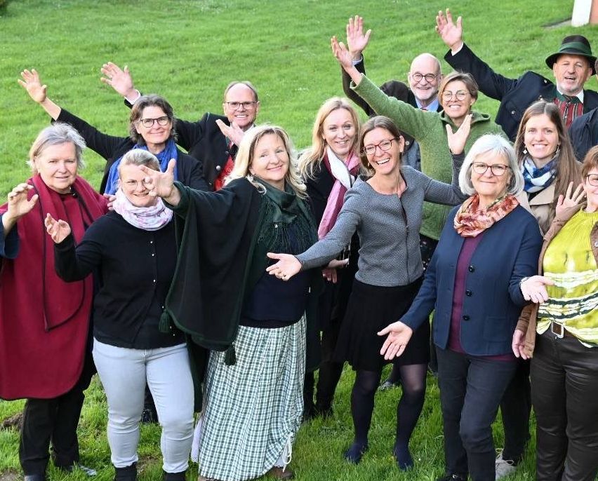 Eine Gruppe von Erwachsenen, darunter Männer und Frauen, stehen auf einer Wiese und lächeln und winken mit ihren Händen, wahrscheinlich für ein Foto.