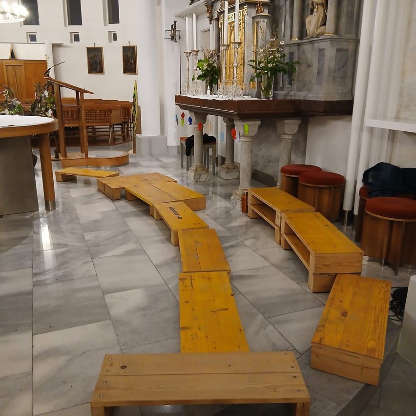Eine Kirche mit hölzernen Bänken und einem mit Blumen geschmückten Altar. In der linken Ecke befindet sich ein Podium mit einem Tisch daneben. Es gibt Stühle und Hocker mit einer Statue hinter dem Altar.