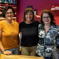 Drei Frauen stehen lächelnd vor einem Tresen und tragen Brillen. Eine trägt ein senfgelbes Shirt, eine ein schwarzes und die andere ein schwarz-weißes Blumenbluse. Hinter ihnen ist eine leuchtend rosa Wand mit verschiedenen Gegenständen.