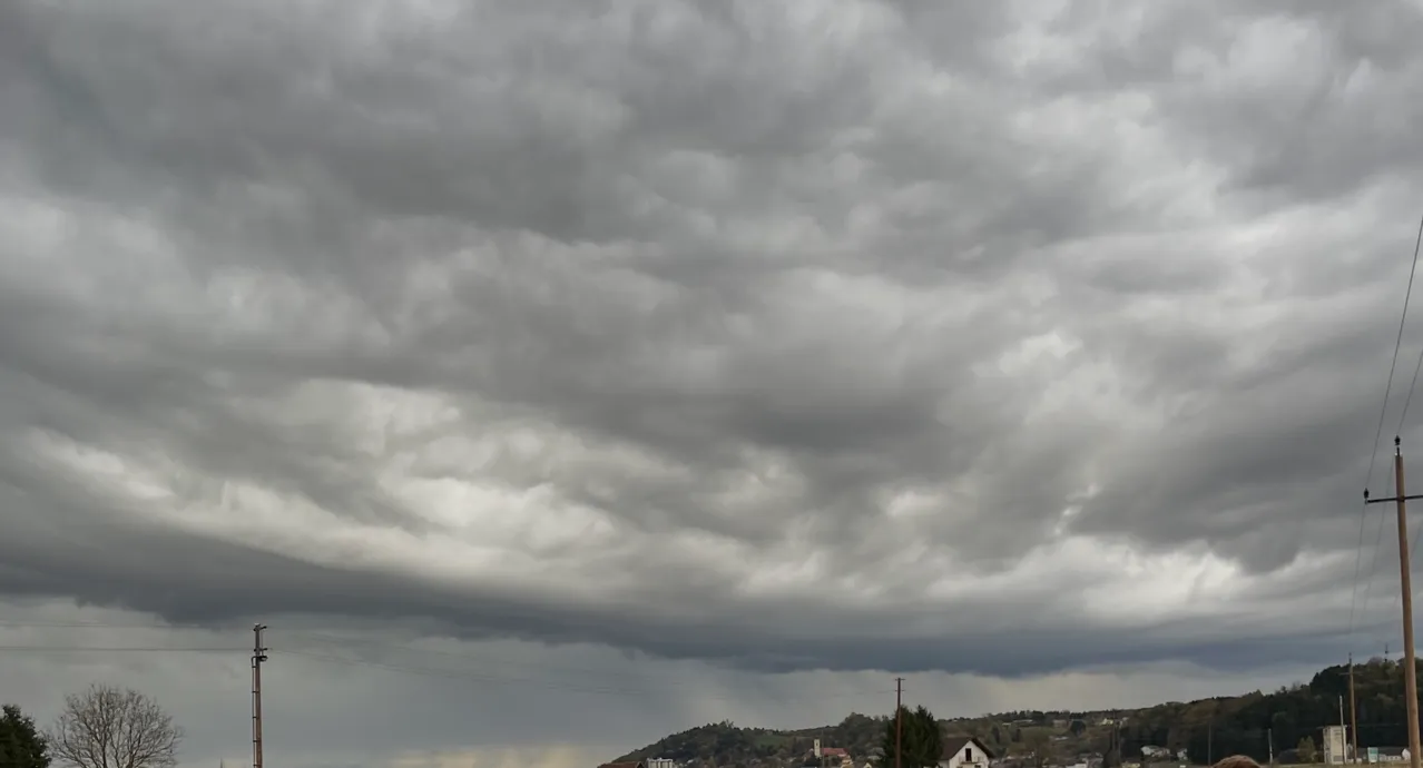 Eine Landschaft unter einem stürmischen Himmel mit dunklen Wolken und Regen. Ein kleines Haus und Bäume sind im Vordergrund sichtbar.