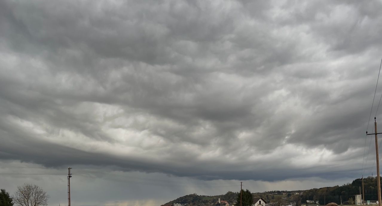 Eine Landschaft unter einem stürmischen Himmel mit dunklen Wolken und Regen. Ein kleines Haus und Bäume sind im Vordergrund sichtbar.