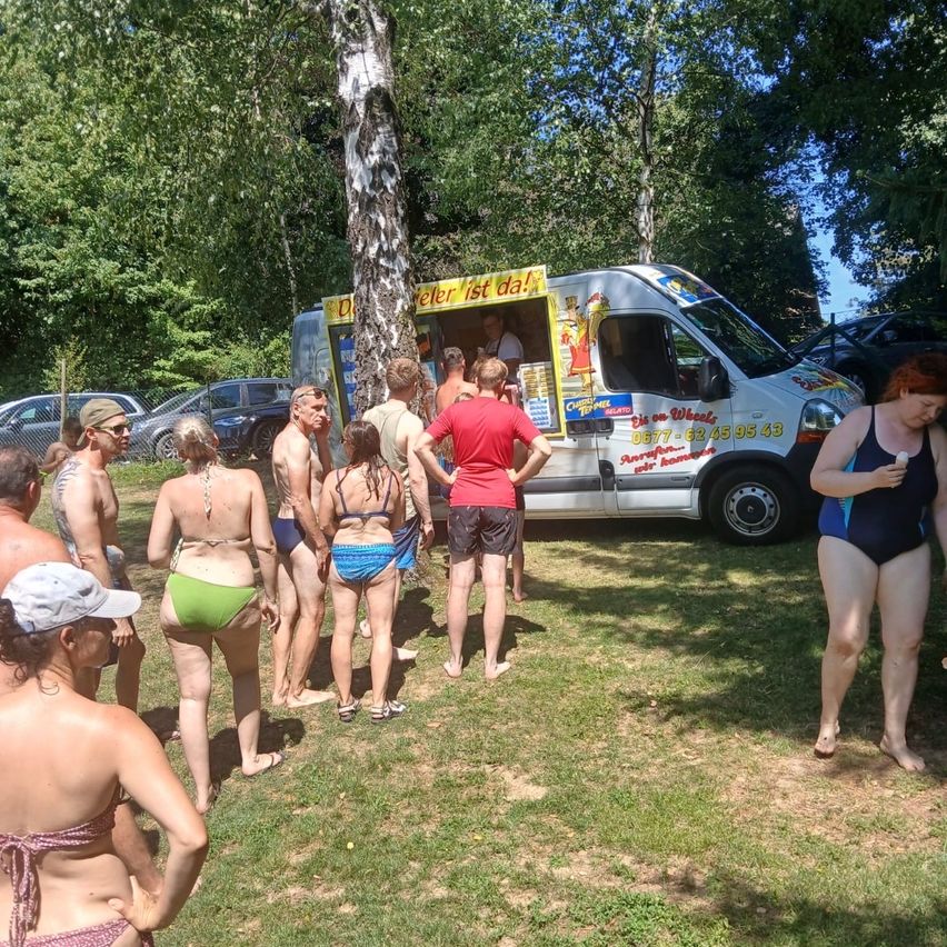 A group of people in swimwear stand near a white ice cream van with a yellow sign, 'Peter ist da!', in a grassy area.