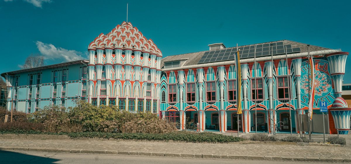 Ein farbenfrohes Gebäude mit Dachturm und Solarmodulen, umgeben von Grün, unter einem klaren blauen Himmel.