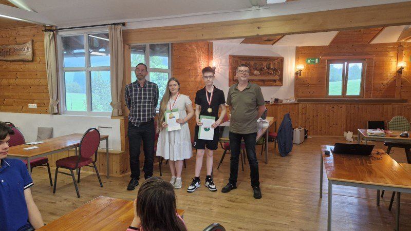 Vier Personen stehen in einem Raum, zwei halten Urkunden, einer eine Medaille. Hinter ihnen steht ein Tisch mit einem Laptop. Der Raum hat Holzwände und -böden, Fenster mit Blick auf eine Landschaft.