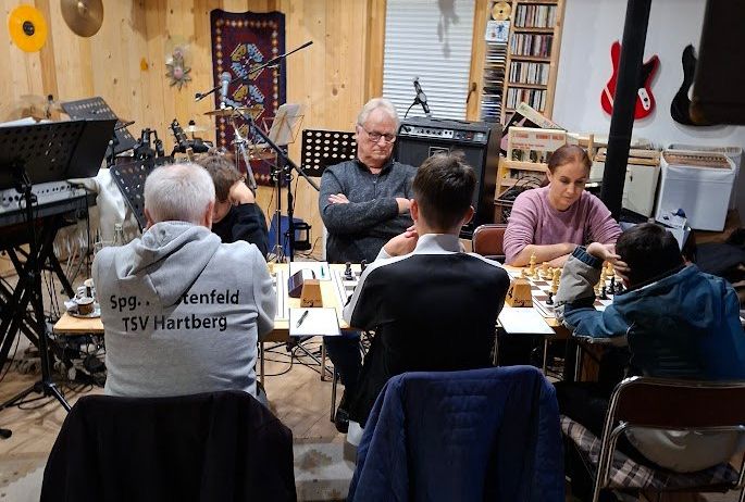 Eine Gruppe von Menschen sitzt um ein Schachbrett herum in einem Raum mit Holzwänden und spielt eine Partie. Ein Mann mit Brille beobachtet sie. Im Hintergrund befinden sich Musikinstrumente und ein Lautsprecher.