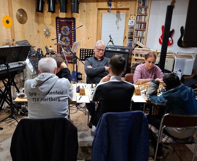 Eine Gruppe von Menschen sitzt um ein Schachbrett herum in einem Raum mit Holzwänden und spielt eine Partie. Ein Mann mit Brille beobachtet sie. Im Hintergrund befinden sich Musikinstrumente und ein Lautsprecher.