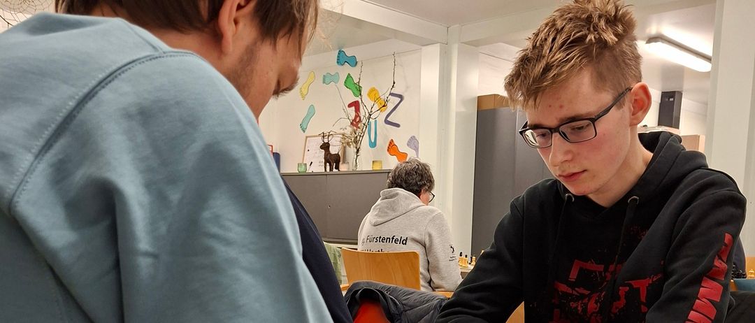 Zwei junge Männer spielen Schach an einem Tisch, einer trägt eine Brille und der andere eine blaue Kapuzenjacke. Das Schachbrett ist mit Figuren aufgebaut, und ein Stift und Papier liegen in der Nähe.