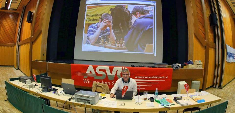 Ein Mann sitzt an einem Tisch mit Laptop vor einer Projektionsleinwand, die Schachspieler zeigt. Der Raum hat Holzwände und ist für eine Schachveranstaltung vorbereitet.