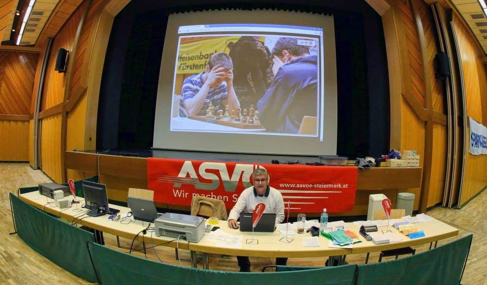 Ein Mann sitzt an einem Tisch mit Laptop vor einer Projektionsleinwand, die Schachspieler zeigt. Der Raum hat Holzwände und ist für eine Schachveranstaltung vorbereitet.