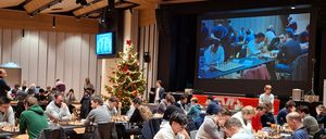 Ein Schachturnier mit Spielern an Tischen. Der Saal hat Holzböden und einen Weihnachtsbaum. Eine große Leinwand zeigt eine Partie.