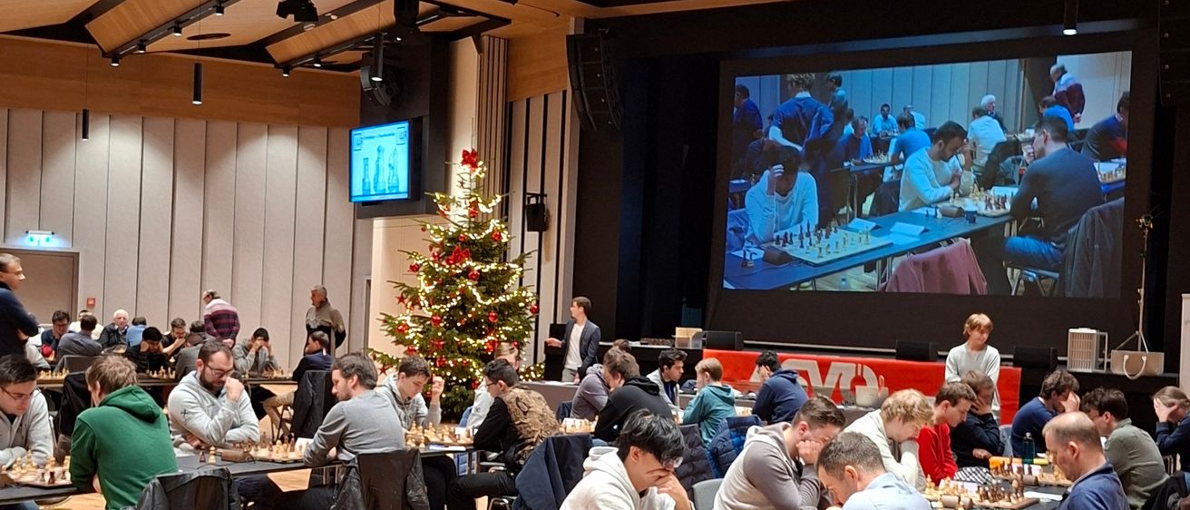 Ein Schachturnier mit Spielern an Tischen. Der Saal hat Holzböden und einen Weihnachtsbaum. Eine große Leinwand zeigt eine Partie.