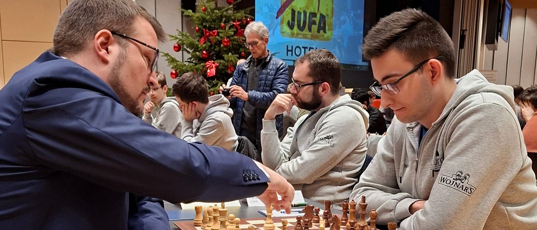 Drei Männer spielen Schach in einem Raum mit einem Weihnachtsbaum, während andere zusehen. Der Mann im blauen Anzug ist kurz davor, zu ziehen.