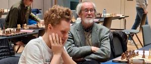 Mehrere Spieler sind in einer Schachpartie engagiert, wobei ein junger Mann im Vordergrund in Gedanken versunken ist. Ein älterer Mann steht hinter ihm mit verschränkten Armen. Der Raum ist gefüllt mit Schachbrettern, Spielern und Beobachtern.