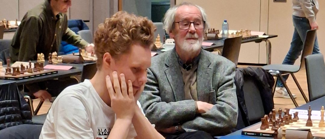 Mehrere Spieler sind in einer Schachpartie engagiert, wobei ein junger Mann im Vordergrund in Gedanken versunken ist. Ein älterer Mann steht hinter ihm mit verschränkten Armen. Der Raum ist gefüllt mit Schachbrettern, Spielern und Beobachtern.