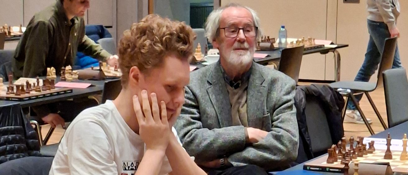 Mehrere Spieler sind in einer Schachpartie engagiert, wobei ein junger Mann im Vordergrund in Gedanken versunken ist. Ein älterer Mann steht hinter ihm mit verschränkten Armen. Der Raum ist gefüllt mit Schachbrettern, Spielern und Beobachtern.