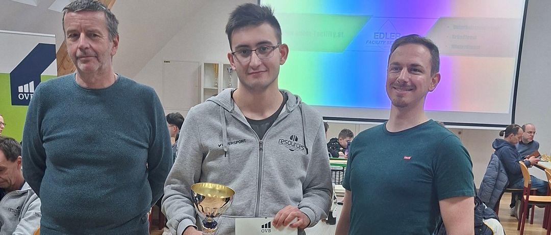 Drei Männer stehen in einem Raum, zwei halten eine Trophäe und ein Zertifikat und lächeln in die Kamera. Hinter ihnen befindet sich eine Projektionsleinwand und einige Leute, die an Tischen sitzen.