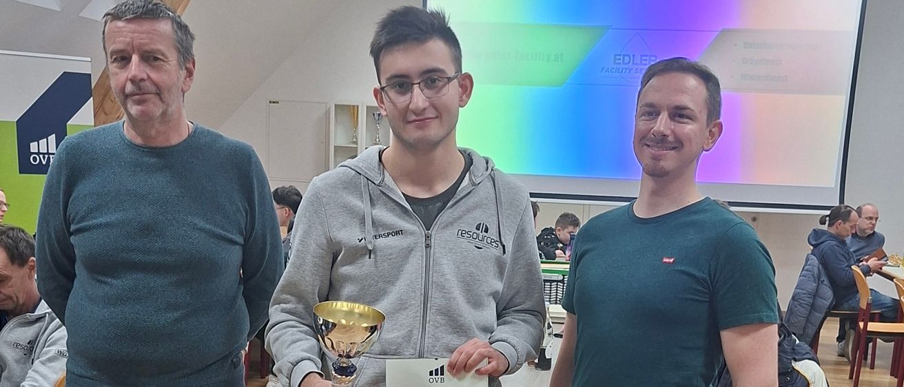 Drei Männer stehen in einem Raum, zwei halten eine Trophäe und ein Zertifikat und lächeln in die Kamera. Hinter ihnen befindet sich eine Projektionsleinwand und einige Leute, die an Tischen sitzen.