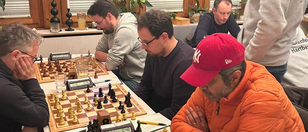 Sechs Männer spielen Schach in einem Raum mit Glasfenstern. Zwei Schachbretter stehen auf dem Tisch. Die Spieler tragen Freizeitkleidung. Ein Mann trägt eine rote Kappe.