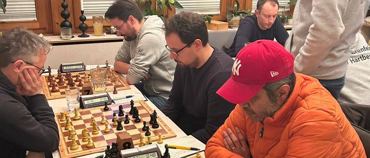 Sechs Männer spielen Schach in einem Raum mit Glasfenstern. Zwei Schachbretter stehen auf dem Tisch. Die Spieler tragen Freizeitkleidung. Ein Mann trägt eine rote Kappe.
