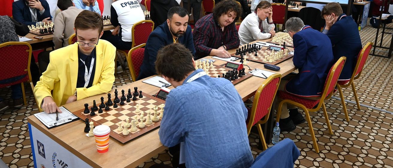 Eine Gruppe von Männern spielt Schach in einem Raum mit Flaggen. Sie sitzen an einem Tisch mit Schachbrettern und Figuren.