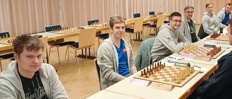 Mehrere junge Männer sitzen um einen langen Tisch herum und spielen Schach. Sie befinden sich in einem Saal mit Holzböden und weißen Vorhängen. Jeder hat ein Schachbrett und Figuren vor sich.