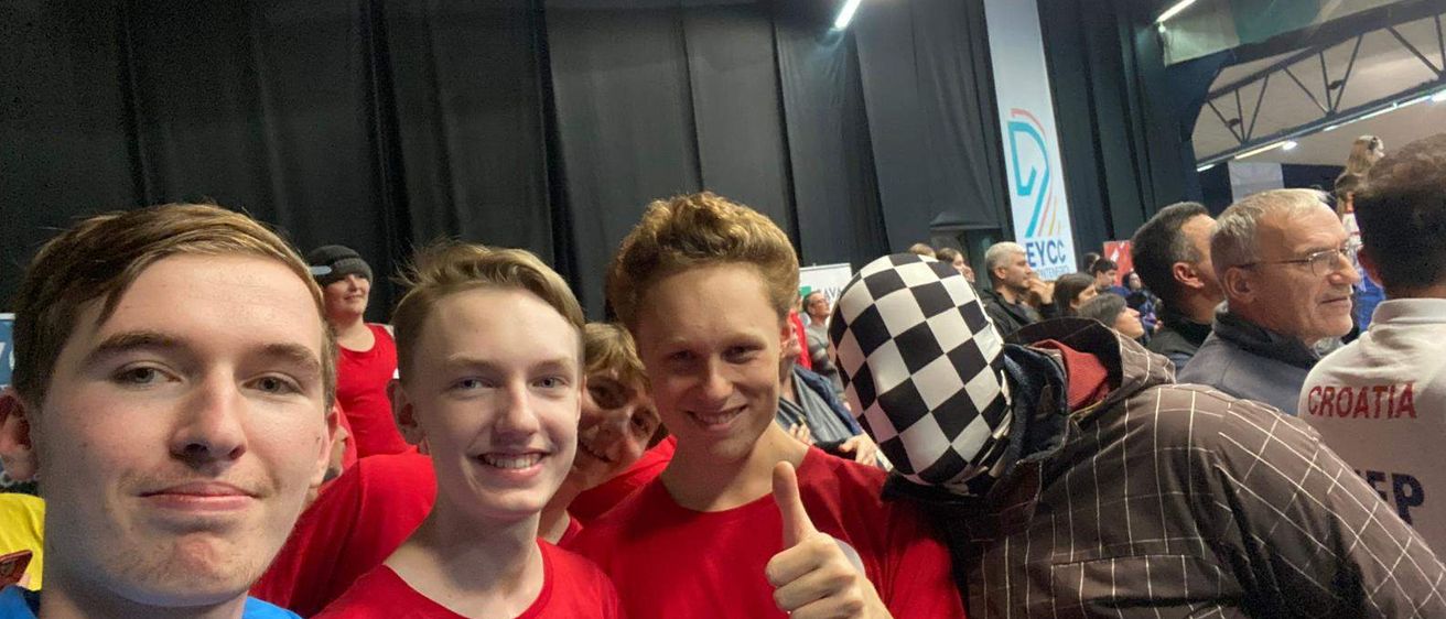 Drei junge Männer in roten Shirts, einer mit Daumen hoch, lächeln zusammen mit einer maskierten Person im Hintergrund.