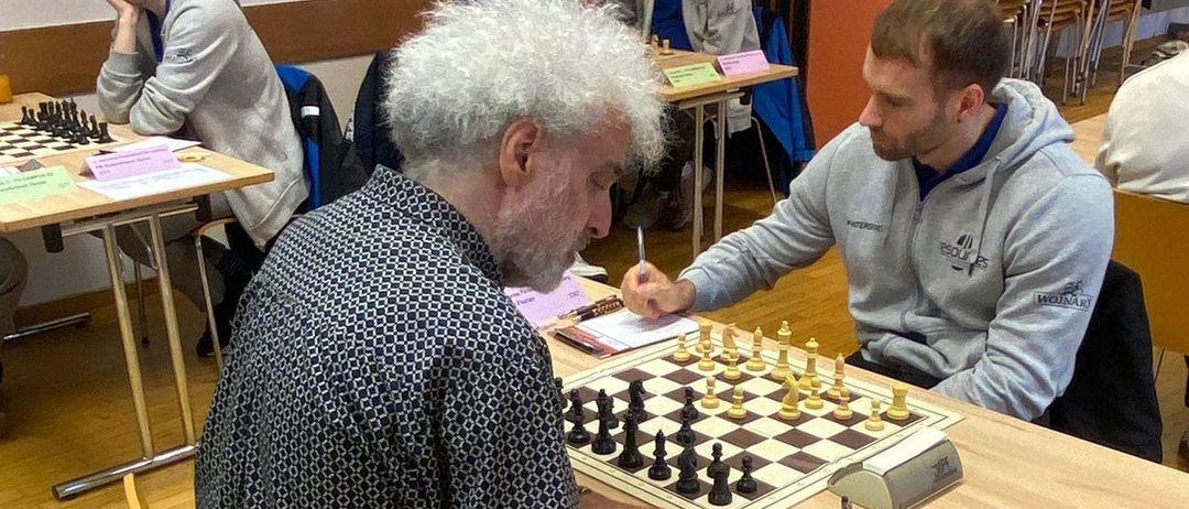 Mehrere Männer spielen Schach in einem Raum. Ein Mann schreibt auf ein Stück Papier. Ein anderer Mann schreibt auf ein Schachbrett.