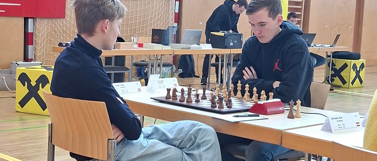 Zwei Jungen spielen Schach in einem Raum mit einem Schachbrett auf dem Tisch. Andere Spieler und Zuschauer sind in der Nähe, einige beschäftigen sich mit anderen Aktivitäten.