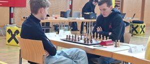 Zwei Jungen spielen Schach in einem Raum mit einem Schachbrett auf dem Tisch. Andere Spieler und Zuschauer sind in der Nähe, einige beschäftigen sich mit anderen Aktivitäten.