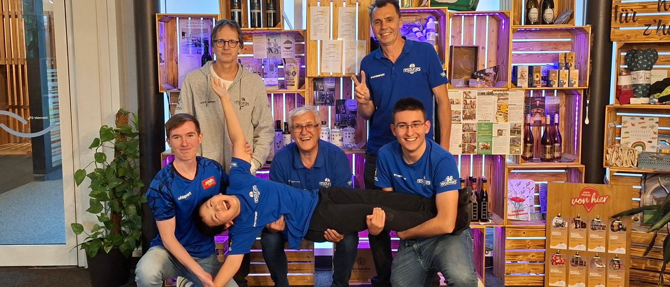 Fünf Männer in blauen Hemden posieren für ein Foto in einem Raum mit Holzregalen, Weinflaschen und einem Schild. Eine Frau macht eine Handstand in der Mitte.