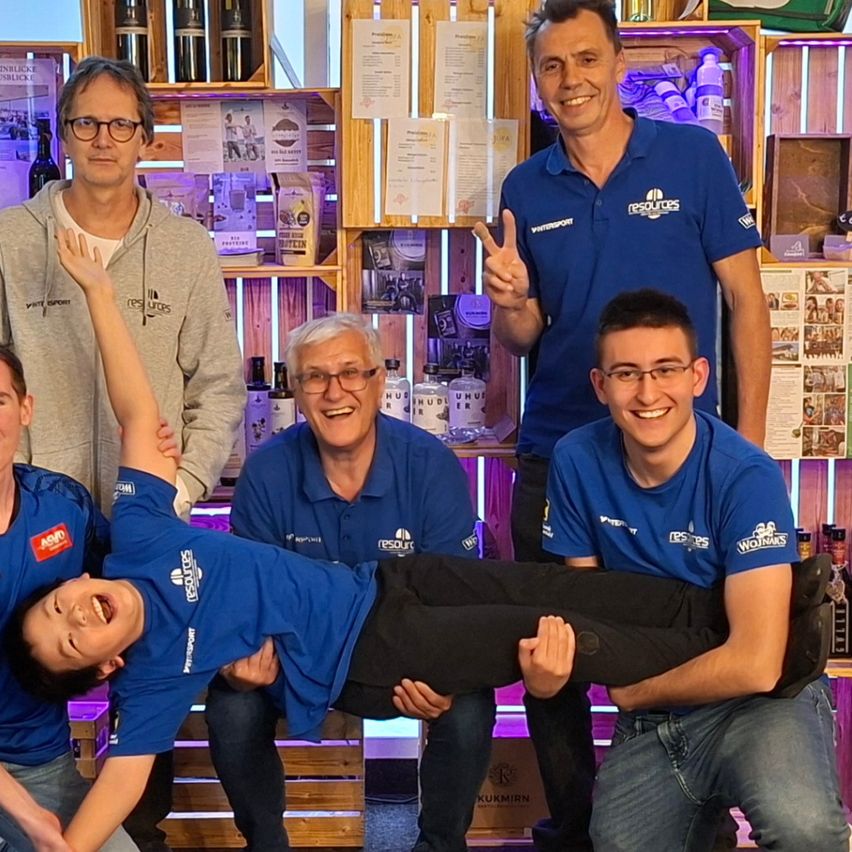 Fünf Männer in blauen Hemden posieren für ein Foto in einem Raum mit Holzregalen, Weinflaschen und einem Schild. Eine Frau macht eine Handstand in der Mitte.
