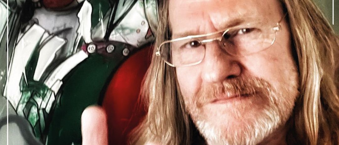 Ein Mann mit langen Haaren und Brille steht vor einer Joker-Wandmalerei. Er macht ein Friedenszeichen. Der Text lautet Kunst von Herbert Wallner.