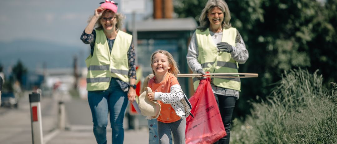 Drei Erwachsene und ein Kind, alle mit Warnwesten bekleidet, gehen auf dem Bürgersteig. Sie tragen Plastiktüten und ein Netz. Das Mädchen hält einen Hut. Sie scheinen die Straße zu reinigen.