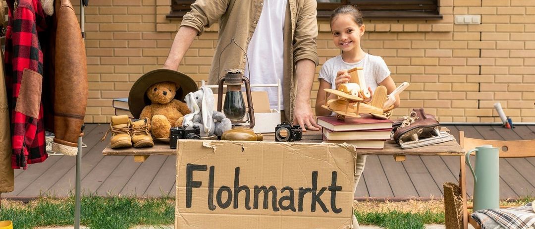 Ein Mann und ein junges Mädchen stehen hinter einem Tisch und verkaufen Gegenstände auf einem Flohmarkt. Verschiedene Artikel befinden sich auf dem Tisch, darunter Bücher, eine Laterne und ein Teddybär.
